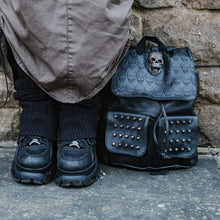 Load image into Gallery viewer, The GothX twin pocket skull vegan backpack sat outside next to a brown brick wall next to a y2k grunge styled model wearing a cargo skirt, leg warmers and black buffalo platform trainers. The vegan leather gothic style bag is facing forward to highlight the diamante effect skull, skull embossed vegan leather front flap, tassel tie cords and two silver studded front pockets.