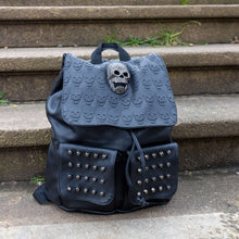 Load image into Gallery viewer, The GothX twin pocket skull vegan backpack sat on a concrete stairway outside. The vegan leather gothic style bag is facing forward to highlight the diamante effect skull, skull embossed vegan leather front flap, tassel tie cords and two silver studded front pockets.