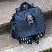 Load image into Gallery viewer, The GothX twin pocket skull vegan backpack sat on a concrete stairway outside. The vegan leather gothic style bag is facing forward to highlight the diamante effect skull, skull embossed vegan leather front flap, tassel tie cords and two silver studded front pockets.