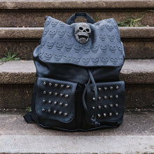 Load image into Gallery viewer, The GothX twin pocket skull vegan backpack sat on a concrete stairway outside. The vegan leather gothic style bag is facing forward to highlight the diamante effect skull, skull embossed vegan leather front flap, tassel tie cords and two silver studded front pockets.