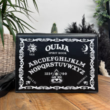 Load image into Gallery viewer, The GothX Ouija Spirit Book Mini Bag sat on a wooden table with green foliage and plants behind. The bag is facing forward to highlight the ouija spirit book white printed detailing on the black book bag with the 3D planchette stitching. Next to the bag on the left is the adjustable detachable strap. Bag is inspired by witchy style and necromancy.
