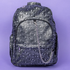 The Rustic Silver Spider Backpack sat on a purple background. The bag is facing forward to highlight the two zip front pockets, two elasticated side pockets, the double zip main compartment with a silver draping chain across the front. The vegan friendly faux leather bag has 3d embossed spiders in varying sizes with brushed black and silver tones all over.