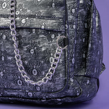 Load image into Gallery viewer, A close up of the Rustic Silver Spider Backpack sat on a purple background. The bag is facing forward to highlight the two zip front pockets, two elasticated side pockets, the double zip main compartment with a silver draping chain across the front. The vegan friendly faux leather bag has 3d embossed spiders in varying sizes with brushed black and silver tones all over.
