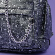 Load image into Gallery viewer, A close up of the Rustic Silver Spider Backpack sat on a purple background. The bag is facing forward to highlight the two zip front pockets, two elasticated side pockets, the double zip main compartment with a silver draping chain across the front. The vegan friendly faux leather bag has 3d embossed spiders in varying sizes with brushed black and silver tones all over.