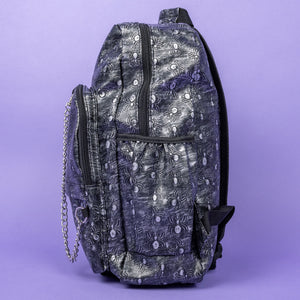 The Rustic Silver Spider Backpack sat on a purple background. The bag is facing left to highlight the two zip front pockets, two elasticated side pockets, the double zip main compartment with a silver draping chain across the front. The vegan friendly faux leather bag has 3d embossed spiders in varying sizes with brushed black and silver tones all over.