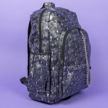 Load image into Gallery viewer, The Rustic Silver Spider Backpack sat on a purple background. The bag is facing right to highlight the two zip front pockets, two elasticated side pockets, the double zip main compartment with a silver draping chain across the front. The vegan friendly faux leather bag has 3d embossed spiders in varying sizes with brushed black and silver tones all over.