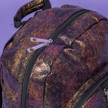 Load image into Gallery viewer, Close up of The Rustic Skulls &amp; Roses Backpack sat on a purple background. The bag is facing forward to highlight the two zip front pockets, top handle, the double zip main compartment with a silver draping chain across the front. The bag is varying colours of bronze, gold, rose and brown with black 3d embossed sugar skulls, roses and swirls all over.
