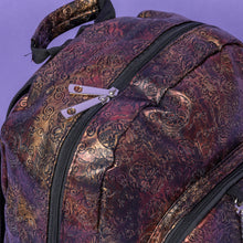 Load image into Gallery viewer, Close up of The Rustic Skulls & Roses Backpack sat on a purple background. The bag is facing forward to highlight the two zip front pockets, top handle, the double zip main compartment with a silver draping chain across the front. The bag is varying colours of bronze, gold, rose and brown with black 3d embossed sugar skulls, roses and swirls all over.