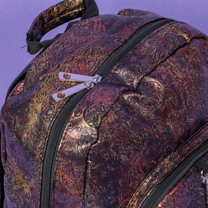 Close up of The Rustic Skulls & Roses Backpack sat on a purple background. The bag is facing forward to highlight the two zip front pockets, top handle, the double zip main compartment with a silver draping chain across the front. The bag is varying colours of bronze, gold, rose and brown with black 3d embossed sugar skulls, roses and swirls all over.