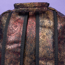 Load image into Gallery viewer, The Rustic Skulls & Roses Backpack sat on a purple background. The bag is facing away from the camera to highlight the top handle and two padded shoulder straps. The bag is varying colours of bronze, gold, rose and brown with black 3d embossed sugar skulls, roses and swirls all over.