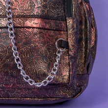 Load image into Gallery viewer, Close up of The Rustic Skulls &amp; Roses Backpack sat on a purple background. The bag is facing forward to highlight the two zip front pockets, two elasticated side pockets, the double zip main compartment with a silver draping chain across the front. The bag is varying colours of bronze, gold, rose and brown with black 3d embossed sugar skulls, roses and swirls all over.
