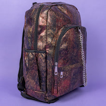 Load image into Gallery viewer, The Rustic Skulls &amp; Roses Backpack sat on a purple background. The bag is facing forward angled right to highlight the two zip front pockets, two elasticated side pockets, the double zip main compartment with a silver draping chain across the front. The bag is varying colours of bronze, gold, rose and brown with black 3d embossed sugar skulls, roses and swirls all over.
