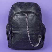 Load image into Gallery viewer, The Black Embossed Cross Backpack sat on a purple background. The backpack is facing forward to highlight the two front silver zip pockets, two elasticated side pockets, main double zip compartment and a silver decorative detachable silver chain draping across the front. The bag is made of vegan friendly leather with 3d embossed crosses in varying sizes all over.