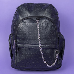 The Black Embossed Cross Backpack sat on a purple background. The backpack is facing forward to highlight the two front silver zip pockets, two elasticated side pockets, main double zip compartment and a silver decorative detachable silver chain draping across the front. The bag is made of vegan friendly leather with 3d embossed crosses in varying sizes all over.