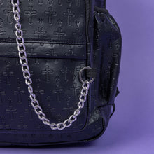 Load image into Gallery viewer, A close up of the Black Embossed Cross Backpack sat on a purple background. The backpack is facing forward to highlight the two front silver zip pockets, two elasticated side pockets, main double zip compartment and a silver decorative detachable silver chain draping across the front. The bag is made of vegan friendly leather with 3d embossed crosses in varying sizes all over.

