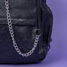 Load image into Gallery viewer, A close up of the Black Embossed Cross Backpack sat on a purple background. The backpack is facing forward to highlight the two front silver zip pockets, two elasticated side pockets, main double zip compartment and a silver decorative detachable silver chain draping across the front. The bag is made of vegan friendly leather with 3d embossed crosses in varying sizes all over.