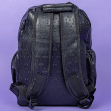 Load image into Gallery viewer, The Black Embossed Cross Backpack sat on a purple background. The backpack is facing away from the camera to highlight the two elasticated side pockets, padded shoulder straps and top handle. The bag is made of vegan friendly leather with 3d embossed crosses in varying sizes all over.