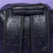 Load image into Gallery viewer, A close up of the Black Embossed Cross Backpack sat on a purple background. The backpack is facing away from the camera to highlight the two elasticated side pockets, padded shoulder straps and top handle. The bag is made of vegan friendly leather with 3d embossed crosses in varying sizes all over.