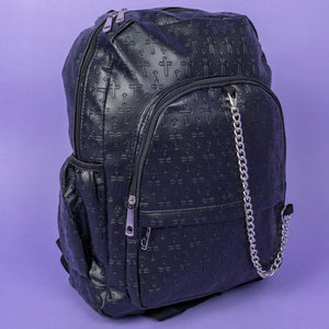 The Black Embossed Cross Backpack sat on a purple background. The backpack is facing forward angled slightly right to highlight the two front silver zip pockets, two elasticated side pockets, main double zip compartment and a silver decorative detachable silver chain draping across the front. The bag is made of vegan friendly leather with 3d embossed crosses in varying sizes all over.