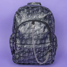 Load image into Gallery viewer, The Rustic Skulls Backpack sat on a purple background. The bag is facing forward to highlight the two front zip pockets, two elasticated side pockets, main zip compartment, the top handle and the detachable decorative silver chain. All over the backpack is an embossed 3d texture skulls and skull and crossbones on a faux leather material in a brushed black and silver grunge style.
