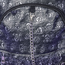 Load image into Gallery viewer, The Rustic Skulls Backpack sat on a purple background. A close up of the bag decorative silver chain detailing across the front. All over the backpack is an embossed 3d texture skulls and skull and crossbones on a faux leather material in a brushed black and silver grunge style.
