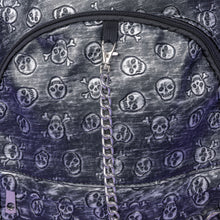 Load image into Gallery viewer, The Rustic Skulls Backpack sat on a purple background. A close up of the bag decorative silver chain detailing across the front. All over the backpack is an embossed 3d texture skulls and skull and crossbones on a faux leather material in a brushed black and silver grunge style.
