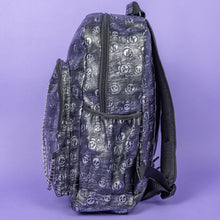 Load image into Gallery viewer, The Rustic Skulls Backpack sat on a purple background. The bag is facing left to highlight the two front zip pockets, two elasticated side pockets, main zip compartment, the top handle and the detachable decorative silver chain. All over the backpack is an embossed 3d texture skulls and skull and crossbones on a faux leather material in a brushed black and silver grunge style.