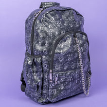Load image into Gallery viewer, The Rustic Skulls Backpack sat on a purple background. The bag is facing forward angled slightly right to highlight the two front zip pockets, two elasticated side pockets, main zip compartment, the top handle and the detachable decorative silver chain. All over the backpack is an embossed 3d texture skulls and skull and crossbones on a faux leather material in a brushed black and silver grunge style.