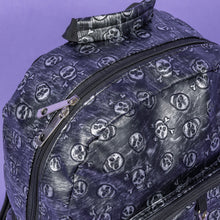 Load image into Gallery viewer, The Rustic Skulls Backpack sat on a purple background. A close up of the bag top handle and double zip opening main compartment. All over the backpack is an embossed 3d texture skulls and skull and crossbones on a faux leather material in a brushed black and silver grunge style.