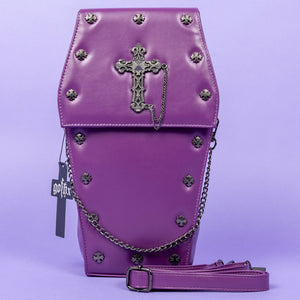 The GothX Purple Coffin Backpack. sat facing forward on a lilac background. The coffin bag in a sleek faux leather is a coffin shape with a magnetic close clip flap with a cross chain emblem in the centre, surrounding the coffin shape is mini metal cross studs. Draping across the front of a silver detachable decorative chain with the GothX black cross tag hanging to the left side.