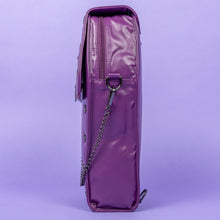 Load image into Gallery viewer, The GothX Purple Coffin Backpack. sat facing left on a lilac background. The coffin bag in a sleek faux leather is a coffin shape with a magnetic close clip flap with a cross chain emblem in the centre, surrounding the coffin shape is mini metal cross studs. Draping across the front of a silver detachable decorative chain with the GothX black cross tag hanging to the left side.
