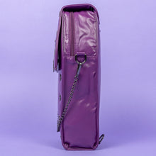 Load image into Gallery viewer, The GothX Purple Coffin Backpack. sat facing left on a lilac background. The coffin bag in a sleek faux leather is a coffin shape with a magnetic close clip flap with a cross chain emblem in the centre, surrounding the coffin shape is mini metal cross studs. Draping across the front of a silver detachable decorative chain with the GothX black cross tag hanging to the left side.