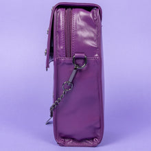 Load image into Gallery viewer, The GothX Purple Mini Coffin Bag sat facing left on a lilac background. The coffin bag in a sleek faux leather is a coffin shape with a magnetic close clip flap with a cross chain emblem in the centre, surrounding the coffin shape is mini metal cross studs. Draping across the front of a silver detachable decorative chain with the GothX black cross tag hanging to the left side.