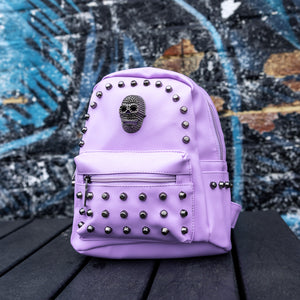  GothX Pastel Lilac Skull Head Small Studs Vegan Mini Backpack sat on a black wooden table in front of a blue graffiti wall. The vegan pastel leather bag is facing forward to highlight the metal stud detailing, the metal skull centrepiece, front zip pocket, two side pockets and top handle. The bag is inspired by pastel goth and soft grunge fashion.