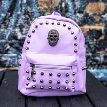 Load image into Gallery viewer, GothX Pastel Lilac Skull Head Small Studs Vegan Mini Backpack sat on a black wooden table in front of a blue graffiti wall. The vegan pastel leather bag is facing forward to highlight the metal stud detailing, the metal skull centrepiece, front zip pocket, two side pockets and top handle. The bag is inspired by pastel goth and soft grunge fashion.
