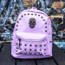 Load image into Gallery viewer, GothX Pastel Lilac Skull Head Small Studs Vegan Mini Backpack sat on a black wooden table in front of a blue graffiti wall. The vegan pastel leather bag is facing forward to highlight the metal stud detailing, the metal skull centrepiece, front zip pocket, two side pockets and top handle. The bag is inspired by pastel goth and soft grunge fashion.