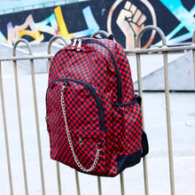Load image into Gallery viewer, The Red Checkerboard Backpack hanging off a metal railing in a skatepark. The vegan friendly bag is facing forward to highlight the red and black check print, two front zip pockets, two elasticated side pockets, main top double zip pocket and silver draping decorative chain.
