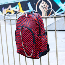 Load image into Gallery viewer, The Red Checkerboard Backpack hanging off a metal railing in a skatepark. The vegan friendly bag is facing forward to highlight the red and black check print, two front zip pockets, two elasticated side pockets, main top double zip pocket and silver draping decorative chain.