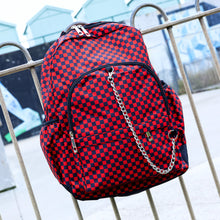 Load image into Gallery viewer, The Red Checkerboard Backpack hanging off a metal railing in a skatepark. The vegan friendly bag is facing forward to highlight the red and black check print, two front zip pockets, two elasticated side pockets, main top double zip pocket and silver draping decorative chain.