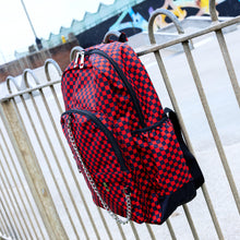 Load image into Gallery viewer, The Red Checkerboard Backpack hanging off a metal railing in a skatepark. The vegan friendly bag is facing forward to highlight the red and black check print, two front zip pockets, two elasticated side pockets, main top double zip pocket and silver draping decorative chain.