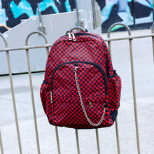 Load image into Gallery viewer, The Red Checkerboard Backpack hanging off a metal railing in a skatepark. The vegan friendly bag is facing forward to highlight the red and black check print, two front zip pockets, two elasticated side pockets, main top double zip pocket and silver draping decorative chain.