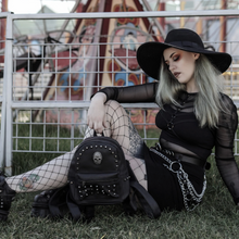 Load image into Gallery viewer, The GothX black vegan skull head mini studded backpack being held by an alternative gothic witchy model with silver hair wearing a gothic styled outfit in front of a fairground ride. The vegan leather bag is facing forward showing off the handle, the skull, mini studs and front zip pocket.
