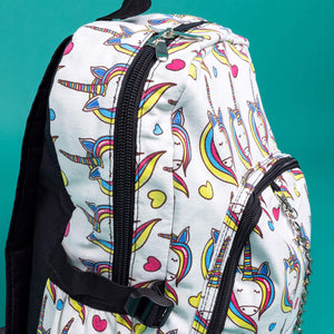 Close up of the Rainbow Unicorn Backpack sat facing right on a teal background. The vegan friendly backpack is a white canvas material with repeating kawaii cute unicorn heads with pink blue and yellow manes with hearts surrounding them. The bag is facing forward to highlight the front two zip pockets, the main zip compartment and silver draping decorative chain.