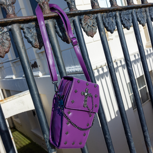 The GothX Purple Mini Coffin Bag hanging off a black metal railing. The coffin bag in a sleek faux leather is a coffin shape with a magnetic close clip flap with a cross chain emblem in the centre, surrounding the coffin shape is mini metal cross studs. Draping across the front of a silver detachable decorative chain.