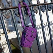 Load image into Gallery viewer, The GothX Purple Mini Coffin Bag hanging off a black metal railing. The coffin bag in a sleek faux leather is a coffin shape with a magnetic close clip flap with a cross chain emblem in the centre, surrounding the coffin shape is mini metal cross studs. Draping across the front of a silver detachable decorative chain.