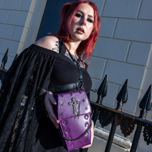 The GothX Purple Mini Coffin Bag being held up by a gothic model wearing a goth black dress with red hair. The coffin bag in a sleek faux leather is a coffin shape with a magnetic close clip flap with a cross chain emblem in the centre, surrounding the coffin shape is mini metal cross studs. Draping across the front of a silver detachable decorative chain with the GothX black cross tag hanging to the left side.