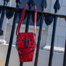 Load image into Gallery viewer, The GothX Red Mini Coffin Bag hanging on a metal black railing outside in Hove, East Sussex. The coffin bag is made with vegan sleek red leather features a detachable metal chain with a large metal cross chain emblem and surrounding cross studs.