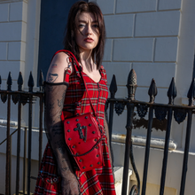 Load image into Gallery viewer, A goth model wearing an emo red tartan dress with the GothX Red Mini Coffin Bag on their shoulder. The coffin bag is made with vegan friendly sleek red leather and features a detachable decorative metal chain with metal cross studding.