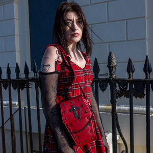 A goth model wearing an emo red tartan dress with the GothX Red Mini Coffin Bag on their shoulder. The coffin bag is made with vegan friendly sleek red leather and features a detachable decorative metal chain with metal cross studding.