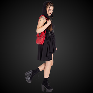 A gothic styled model with dark vampy red makeup wearing a goth black hooded dress holding the GothX Red Mini Coffin Bag on their shoulder with the detachable adjustable strap and walking off to the right on a black background. The coffin bag is made with vegan friendly sleek red leather and features a detachable decorative metal chain with metal cross studding.